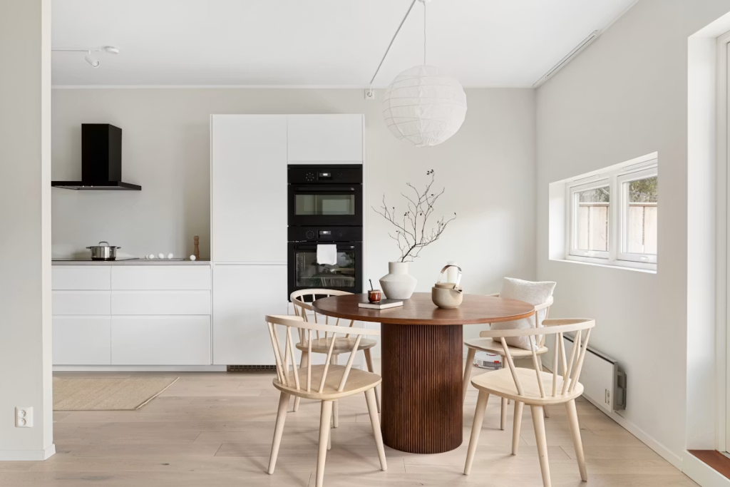 Dark walnut round dining table with a fluted pillar base paired with light wood chairs in a minimalist white kitchen.
