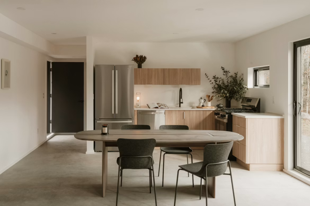 Grey wood oval dining table with minimalist black chairs set in a warm wooden kitchen with a sliding door.