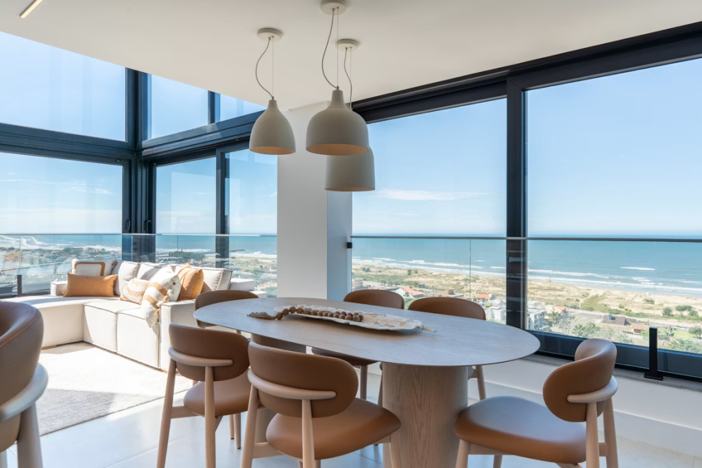 Light ash oval dining table paired with tan leather chairs against high windows overlooking the ocean.