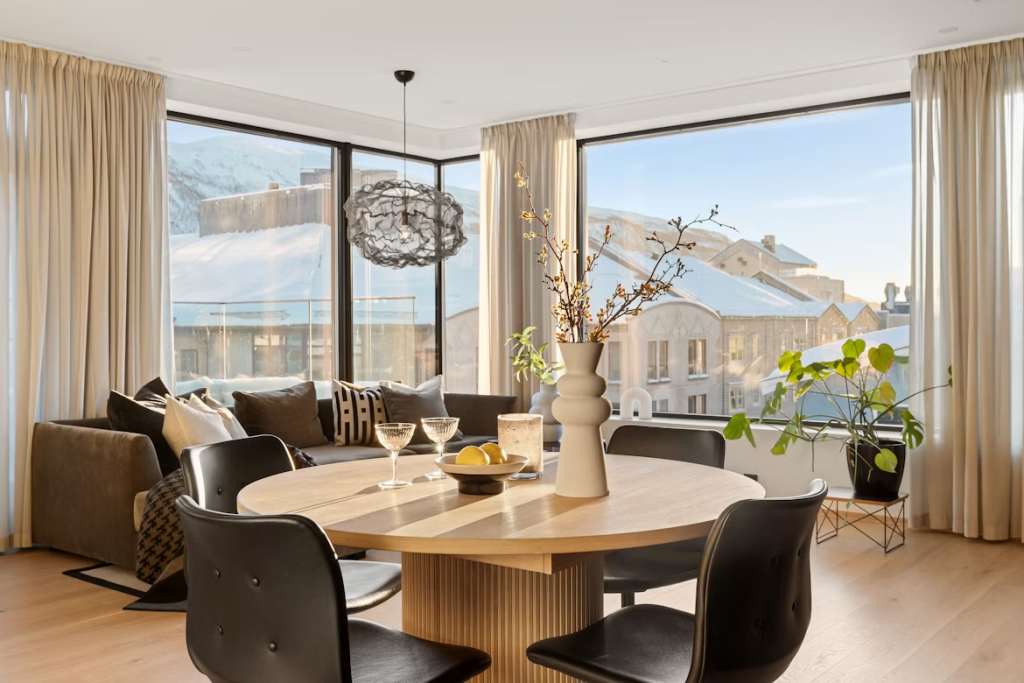 Light oak pedestal round table with black leather chairs set against large windows overlooking a snowy landscape.