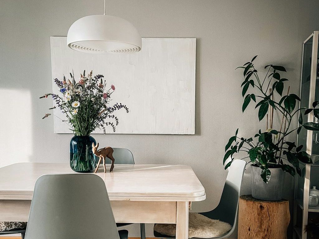 Wildflower bouquet in a dark glass vase on a light wood dining table with modern chairs and a large indoor plant.