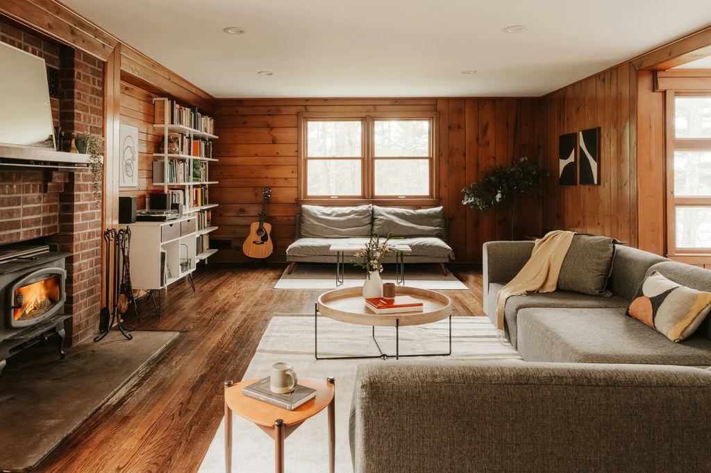 Gray sectional sofa in a wood-paneled living room with a fireplace, coffee table, bookshelves, and large windows.