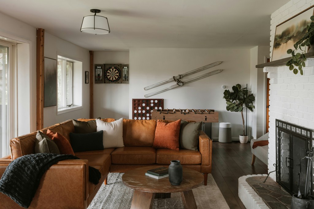 Brown leather sectional sofa with rust and neutral pillows in a cozy living room with a fireplace, coffee table, plants, and game area.