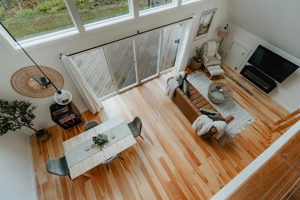 Open tiny home interior with high windows, dining table, leather sofa, lounge chair, and light wood floors