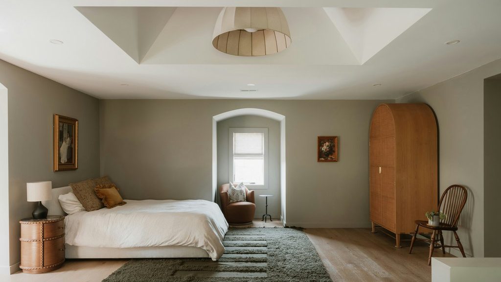 Neutral bedroom with muted sage walls, white bedding, natural wood furniture, and a dark green area rug.