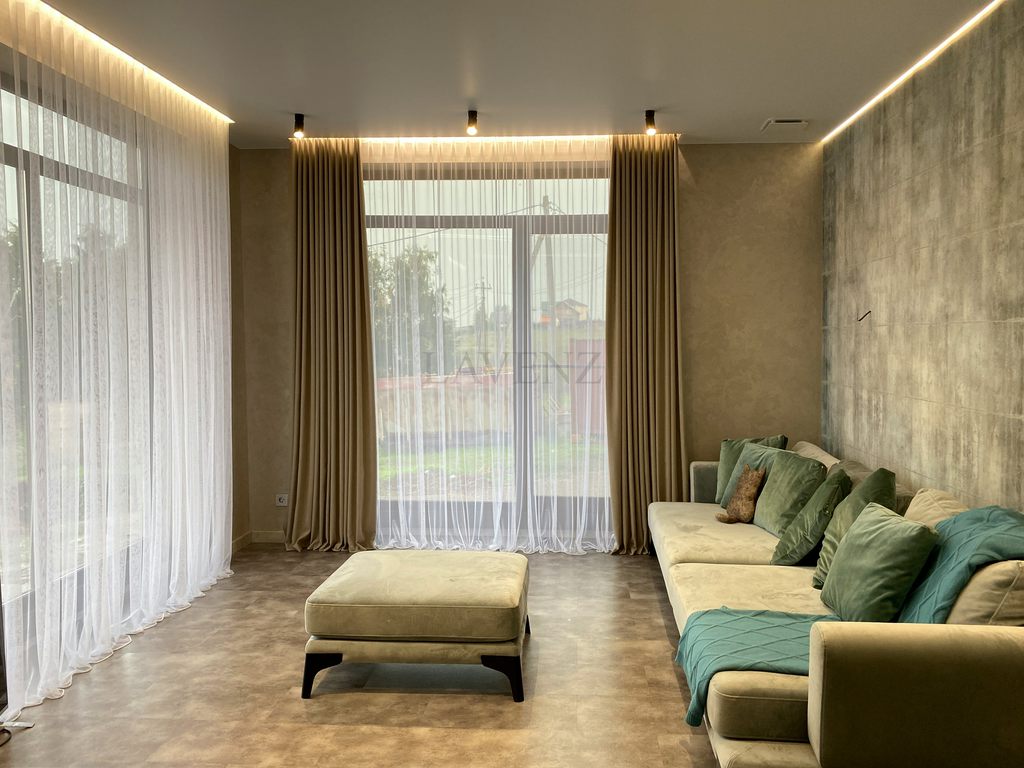 Beige sofa with green cushions and a matching ottoman in a modern living room with floor-to-ceiling windows.