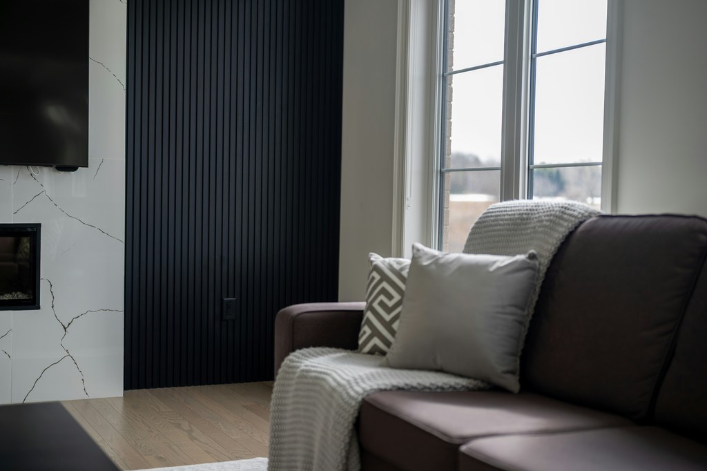 Brown leather sofa with pillows and a throw beside tall windows, a wall-mounted TV, and a fireplace in a modern living room.