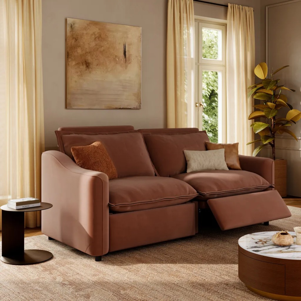 Brown reclining loveseat with one footrest extended in a warm living room with cream curtains, accent pillows, and a round coffee table.