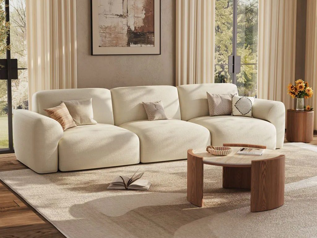 Cream curved sectional sofa with neutral pillows, a light area rug, wood coffee table, and tall windows with beige curtains.