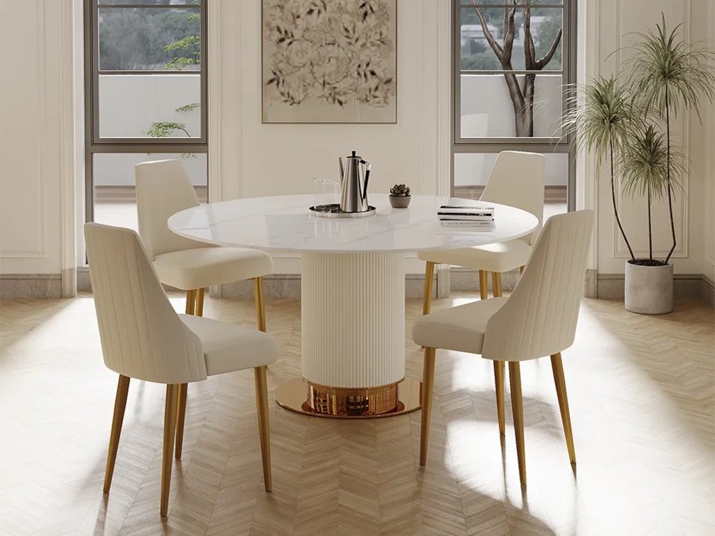 Bright white and gold dining room with a round marble-style table, upholstered cream chairs, gold chair legs, arched windows, and light wood floors.