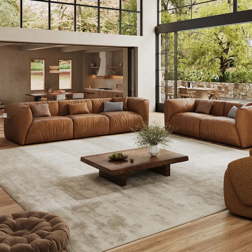 Two caramel leather sofas arranged around a low wood coffee table in a spacious modern living room with floor-to-ceiling windows