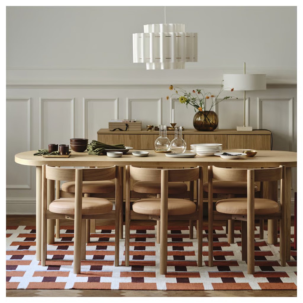 Oval wooden designer dining table with six matching chairs, tableware, and a pendant light in a warm modern dining room.