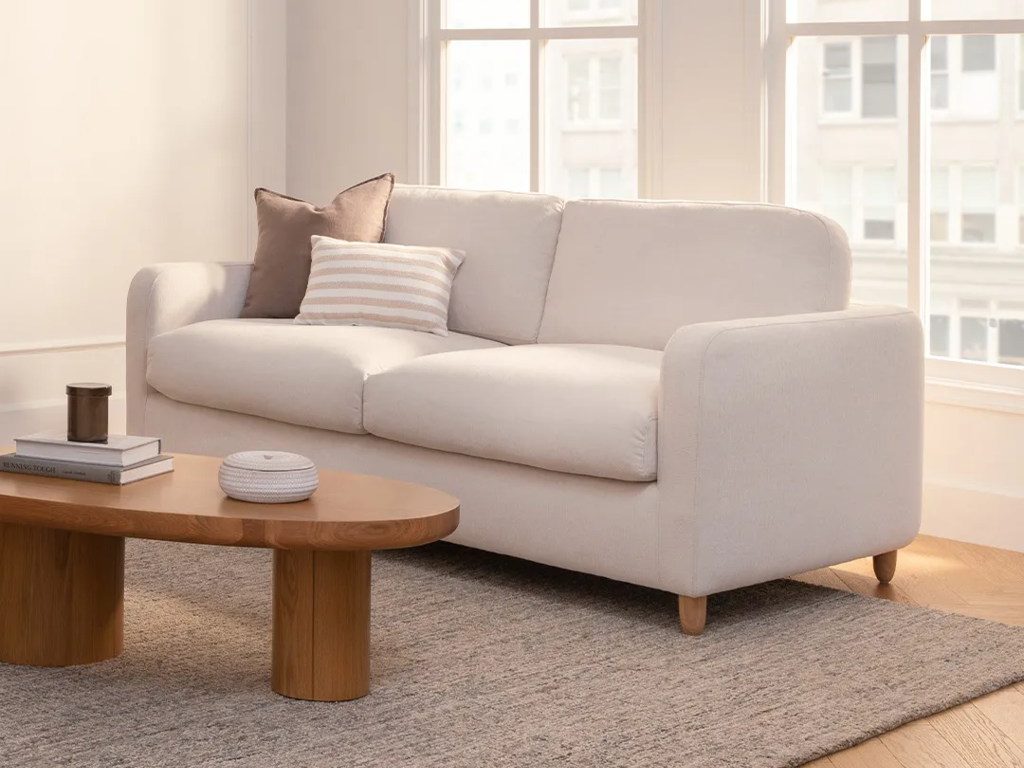 Cream upholstered sleeper sofa with rounded arms and wood legs in a sunlit contemporary living room with a sculptural wood coffee table.
