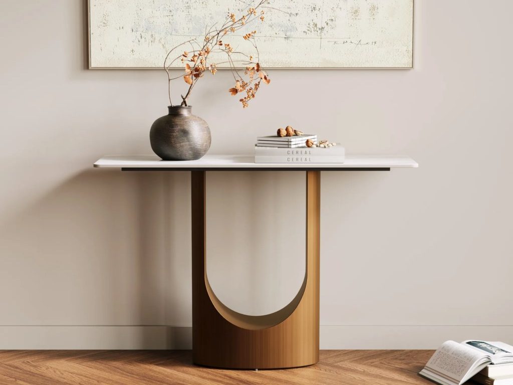 Modern console table with a white tabletop, sculptural gold base, ceramic vase, abstract wall art, wood floor, and neutral decor.