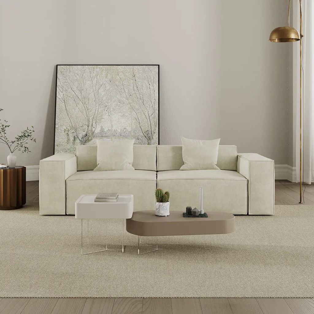 Cream sofa bed styled with white pillows, nesting coffee tables, a floor lamp, and large wall art in a neutral modern living room.