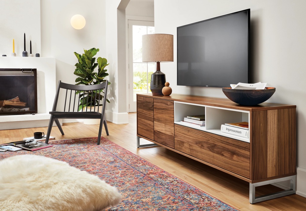 Walnut narrow TV stand with open shelves and closed storage beneath a wall-mounted TV in a bright living room.