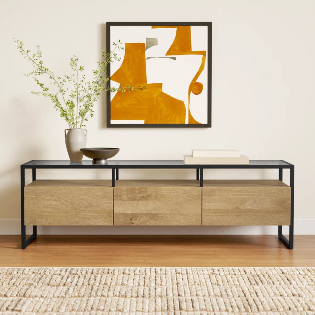 Wood and black metal narrow TV stand with three drawers, open shelves, and framed abstract artwork.
