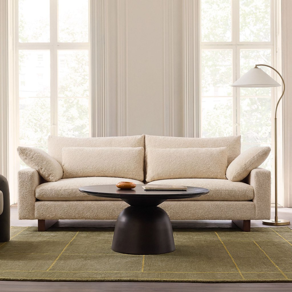 Cream boucle loveseat with loose cushions in a bright living room with large windows and a black coffee table.