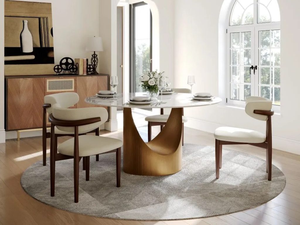 Round pedestal dining table with four upholstered wood chairs in a bright modern dining room with arched windows and a circular rug.