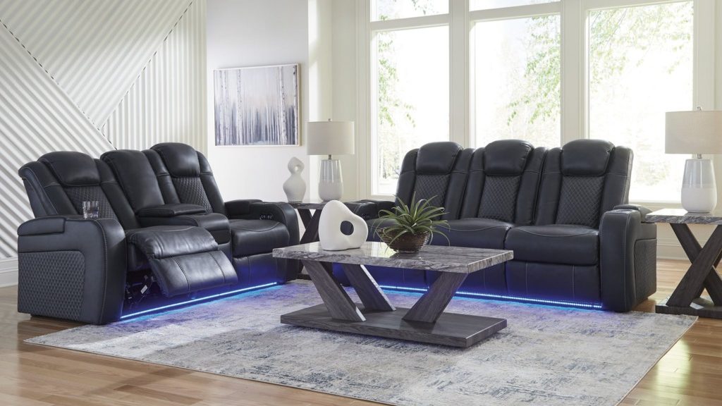 Black power reclining sofa and loveseat set with blue LED base lighting, cupholders, and an extended footrest in a bright modern living room.