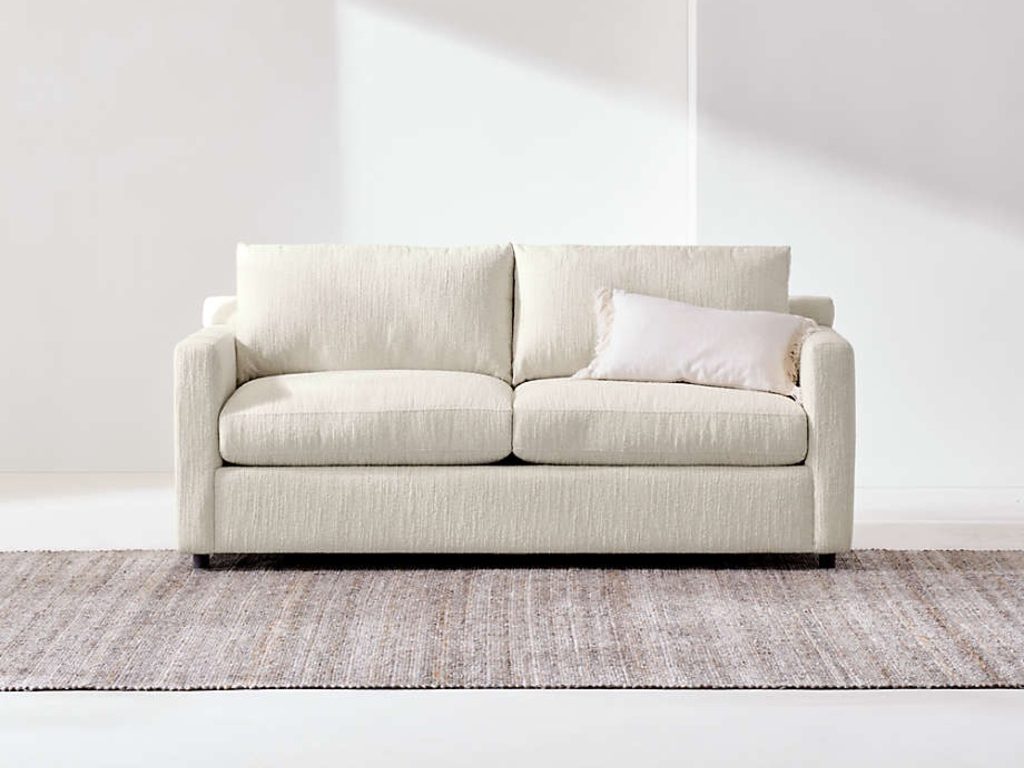 Light beige sleeper sofa with two seat cushions and tapered dark wood legs on a textured rug against a white wall.
