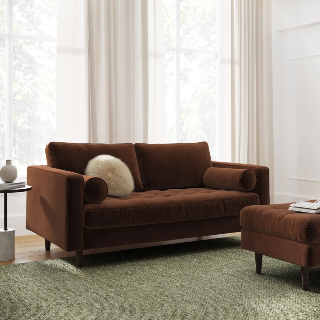 Brown velvet loveseat with tufted seat, bolster pillows, and matching ottoman in a bright modern room.