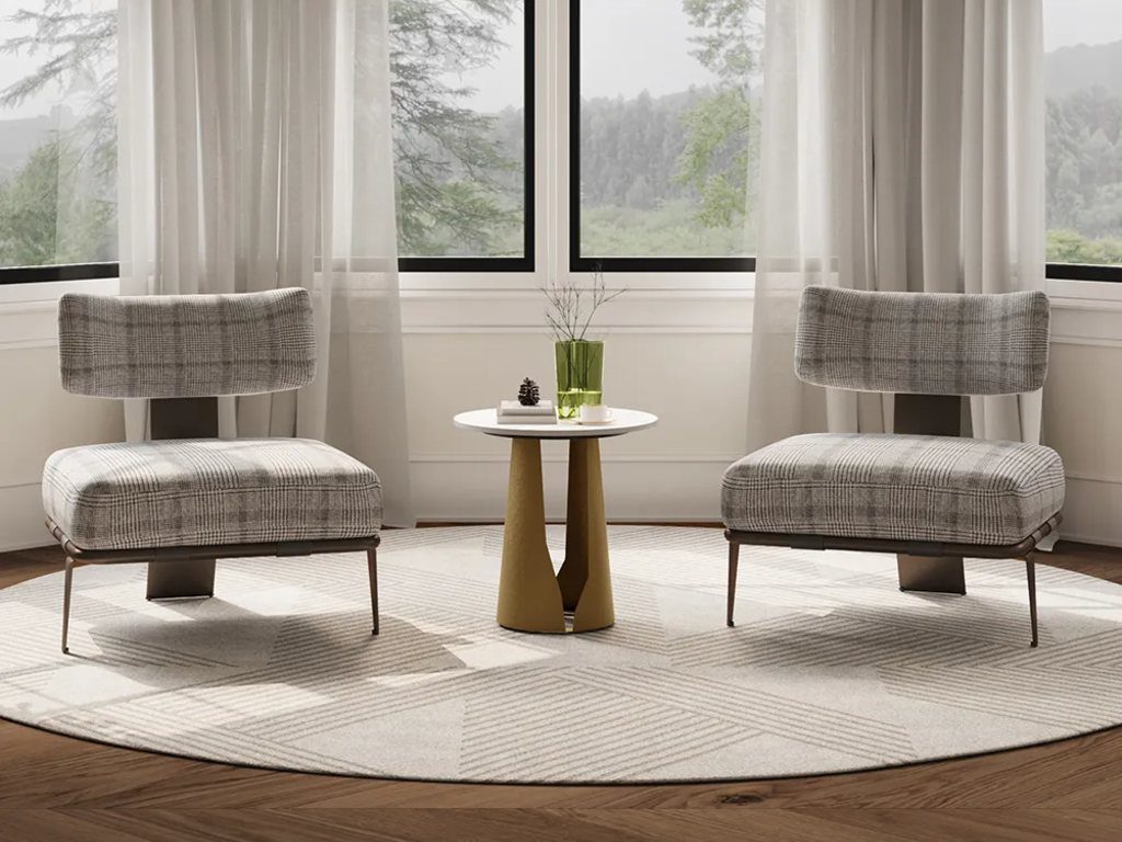 Bright sitting area with two gray plaid accent chairs, a small gold pedestal side table, large black-framed windows, sheer curtains, and a neutral rug.