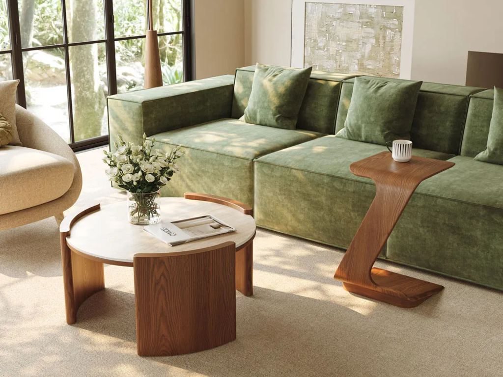 Bright living room with a green sectional, sculptural wood side table, and round white coffee table styled with flowers and a magazine.