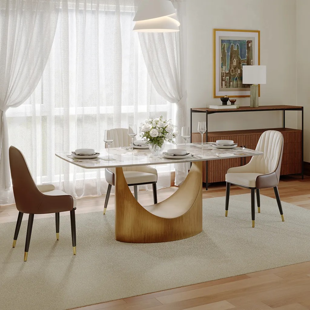 Rectangular designer dining table with a marble-look top, sculptural gold pedestal base, and four upholstered chairs in a bright modern dining room.