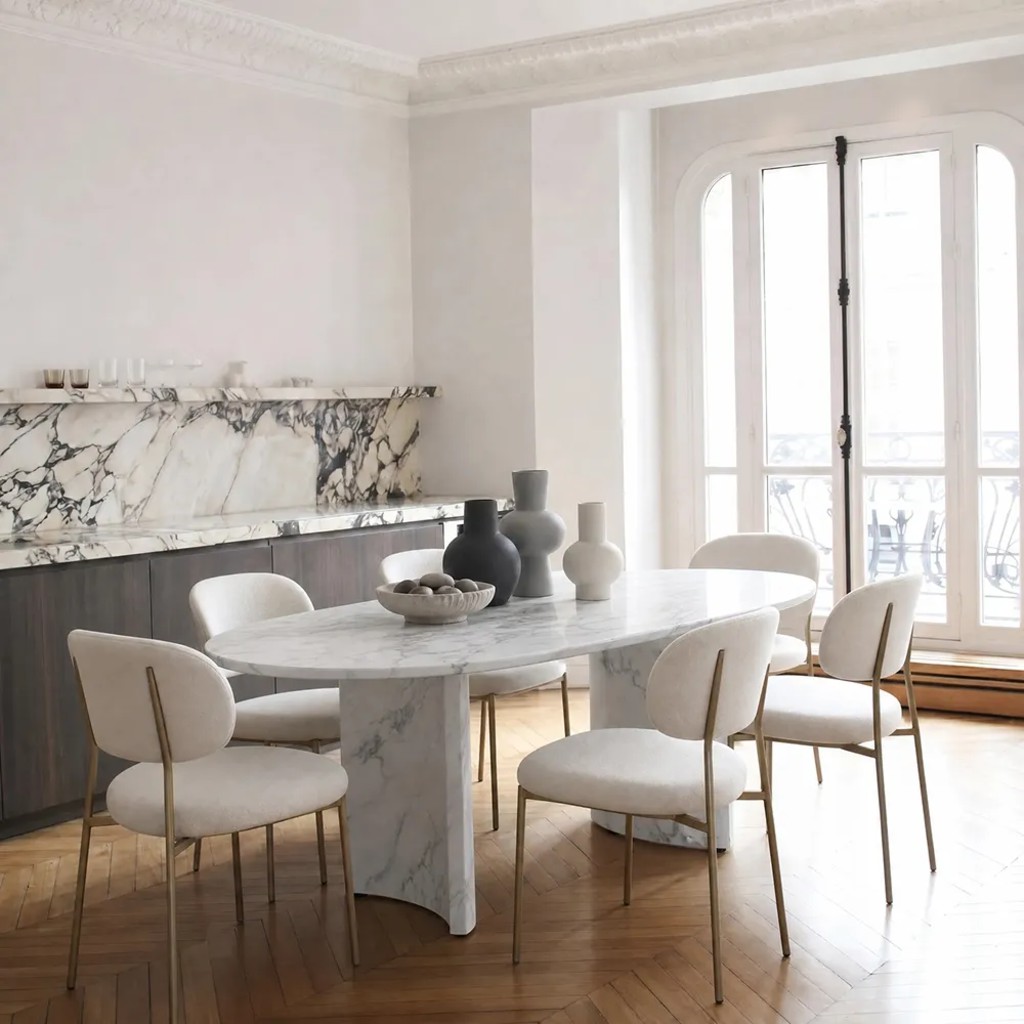 Oval marble designer dining table with a double pedestal base and six upholstered chairs in a bright contemporary dining room.