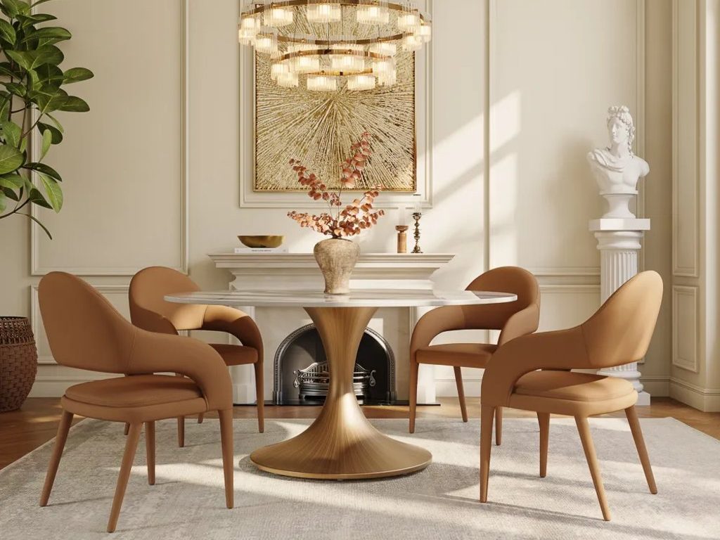 Round pedestal dining table with four tan upholstered chairs in an elegant dining room with a modern chandelier and fireplace.