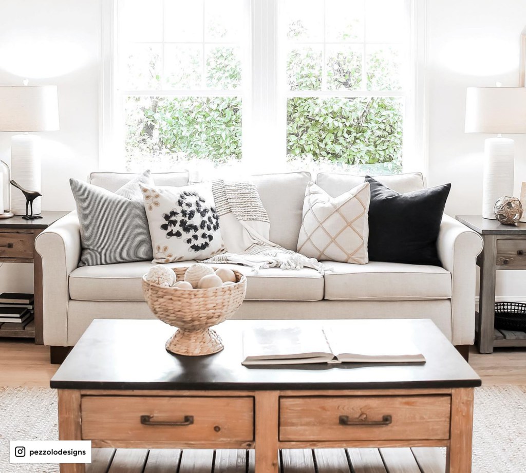White sofa with layered decorative pillows in a bright living room with a wooden coffee table.