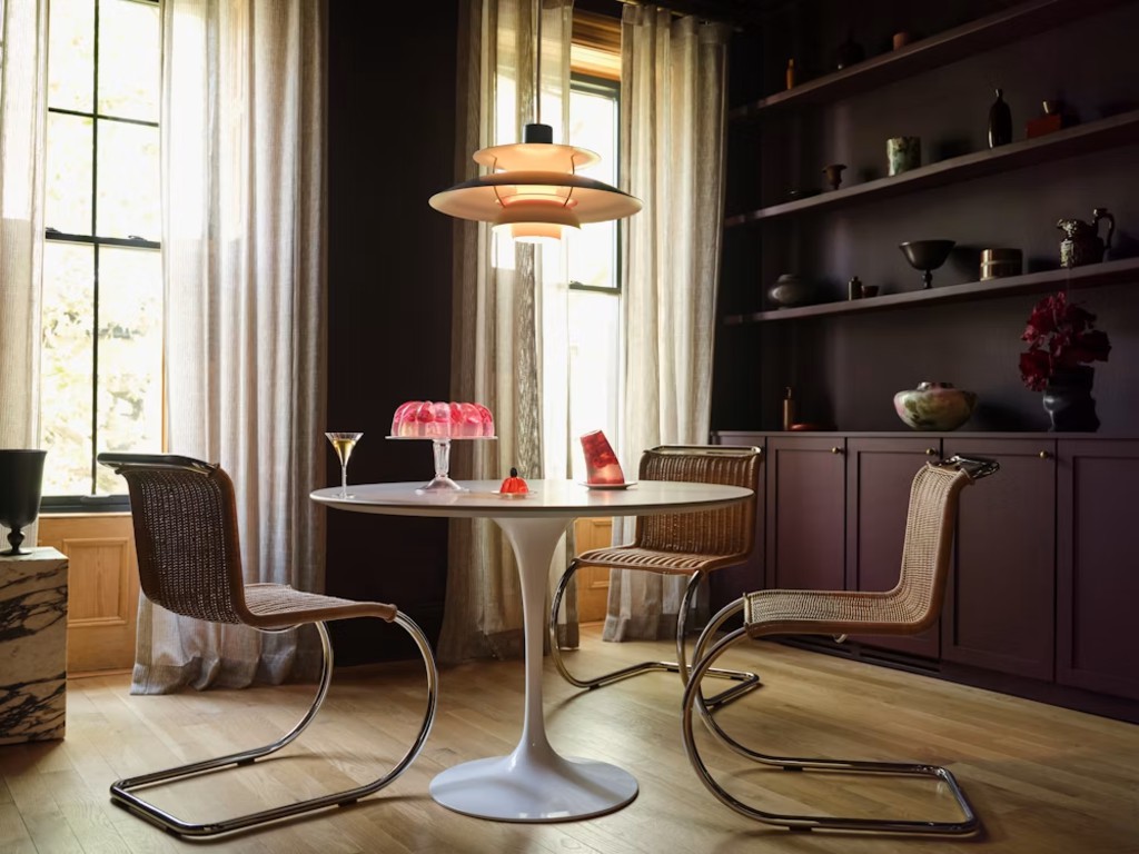 Round white pedestal dining table with three woven cantilever chairs in a dark modern dining room.