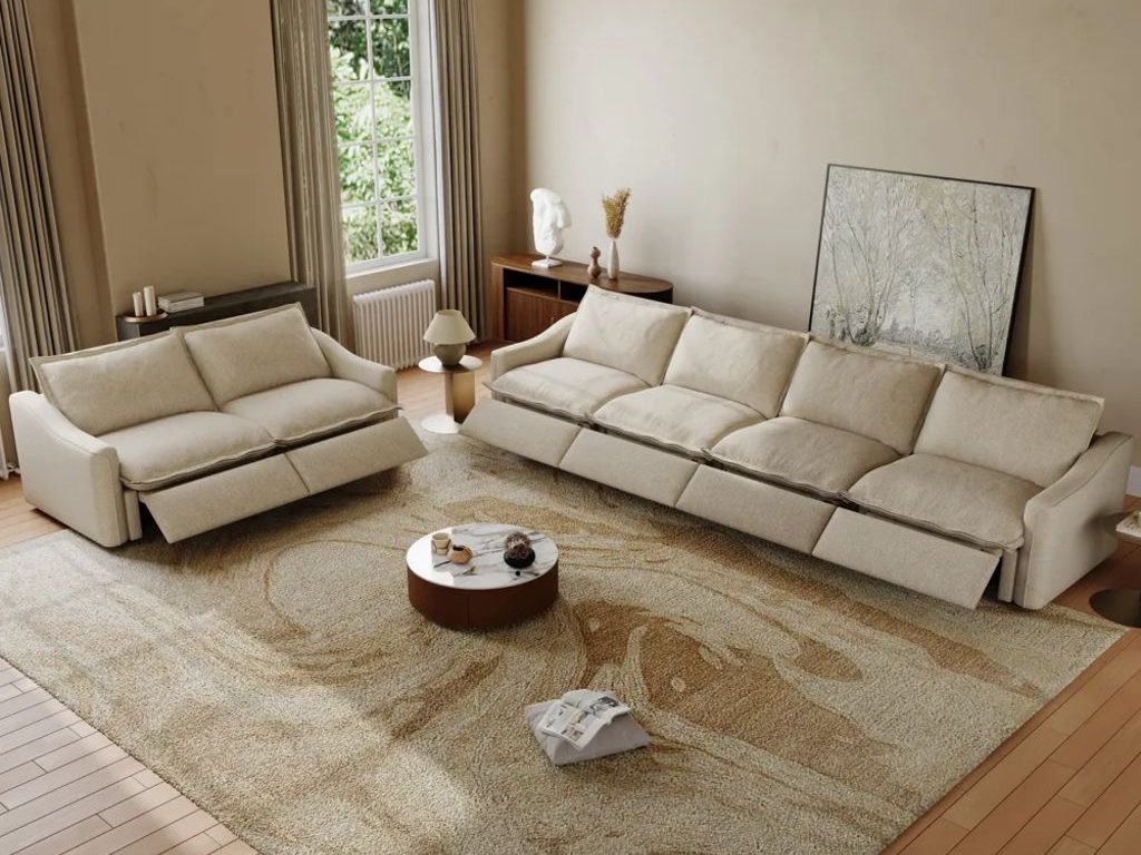 Neutral living room with a cream reclining sofa and loveseat, both footrests extended, arranged around a round coffee table on a large patterned rug.
