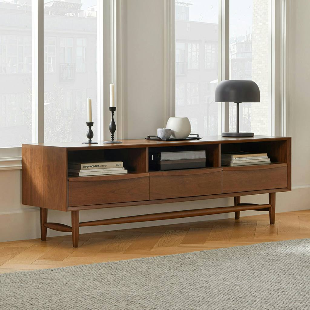 Mid-century walnut TV stand with open shelves and three drawers, styled with books, candles, and a black table lamp in a bright minimalist room.