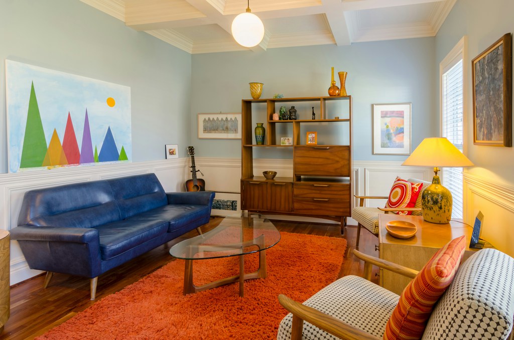 Mid-century modern living room with a blue leather sofa, patterned armchairs, a glass coffee table, warm wood storage, and an orange area rug.