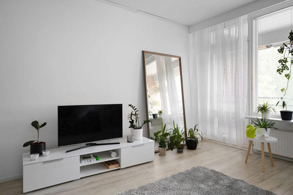 White narrow TV stand with a flat-screen TV in a bright living room with plants and a floor mirror.