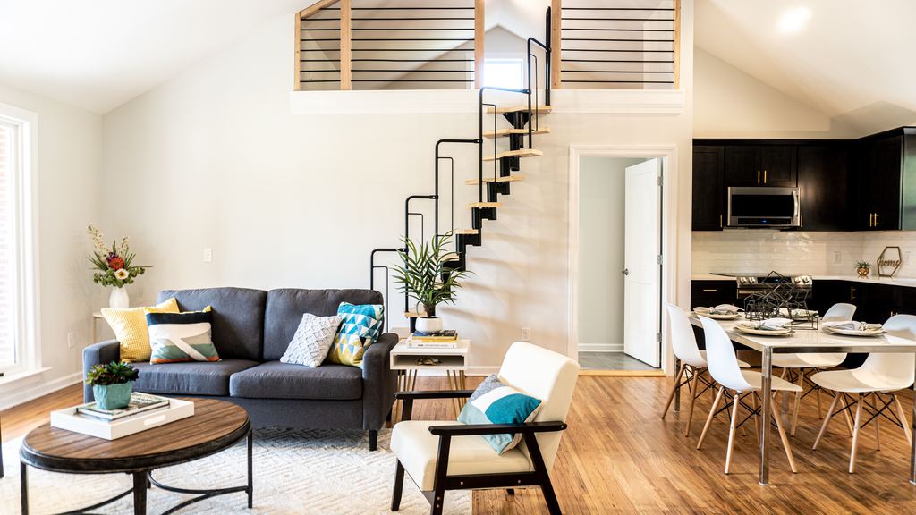 Tiny home interior with loft stairs, gray sofa, round coffee table, dining area, black kitchen cabinets, and light wood floors