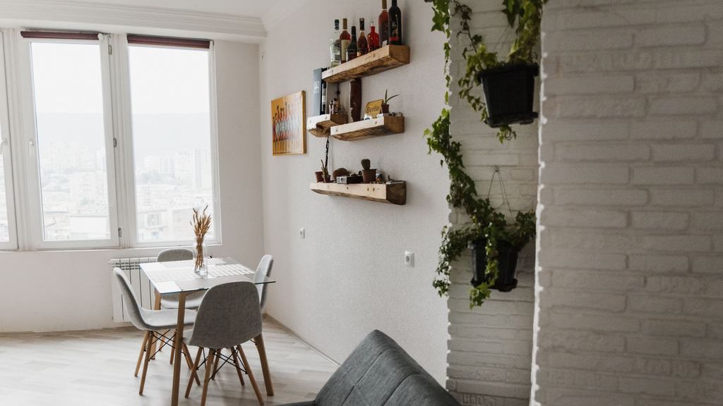 Gray sofa beside rustic floating shelves, trailing plants, and a bright dining area with large windows.