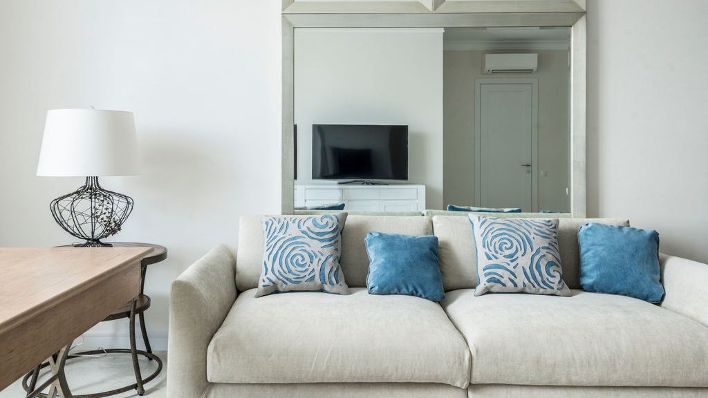 Light beige sofa styled with blue patterned pillows beside a wood table and decorative lamp in a bright modern living room.