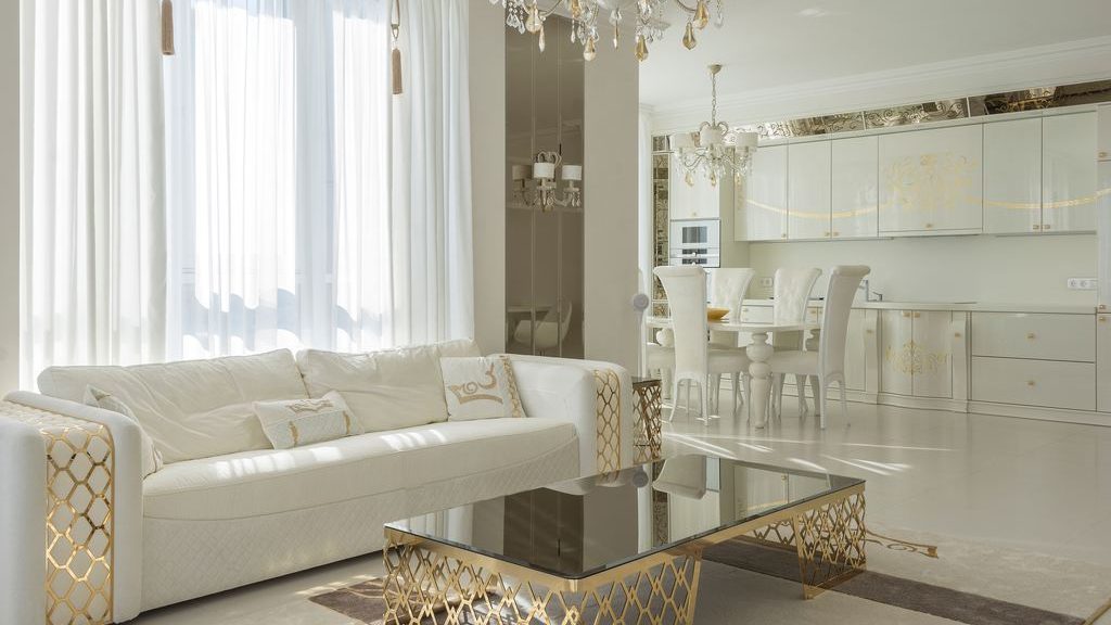 Bright white and gold open-plan living room with a white sofa, glass coffee table, gold geometric accents, chandeliers, and matching dining area.