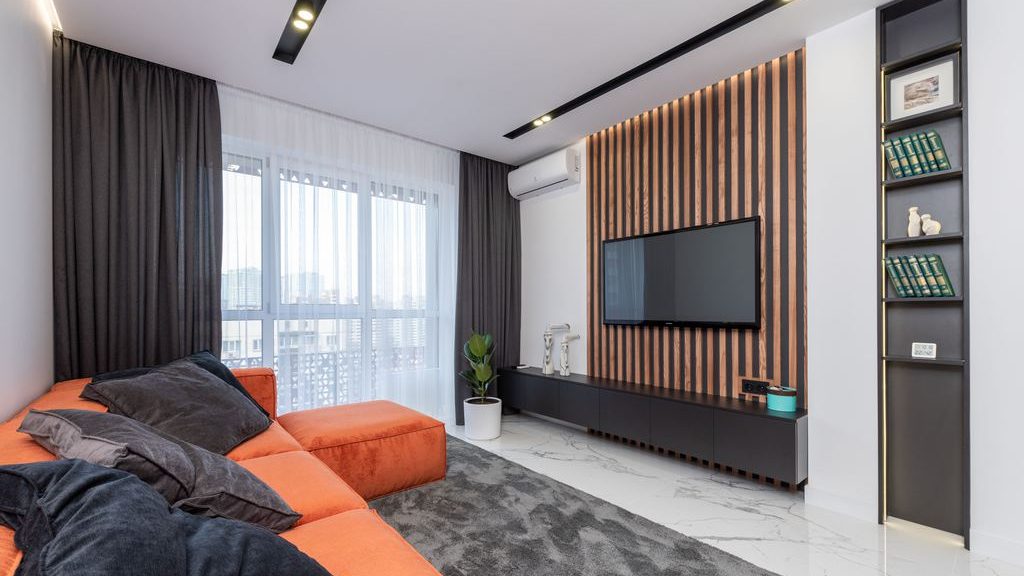 Wall-mounted TV on a vertical wood slat accent wall above a black media console, with an orange sectional sofa, dark rug, built-in shelving, and floor-to-ceiling curtains in a modern living room.