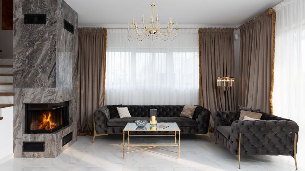 Modern living room with dark tufted sofas, gold metal legs, glass coffee table, chandelier, marble fireplace, and taupe curtains.