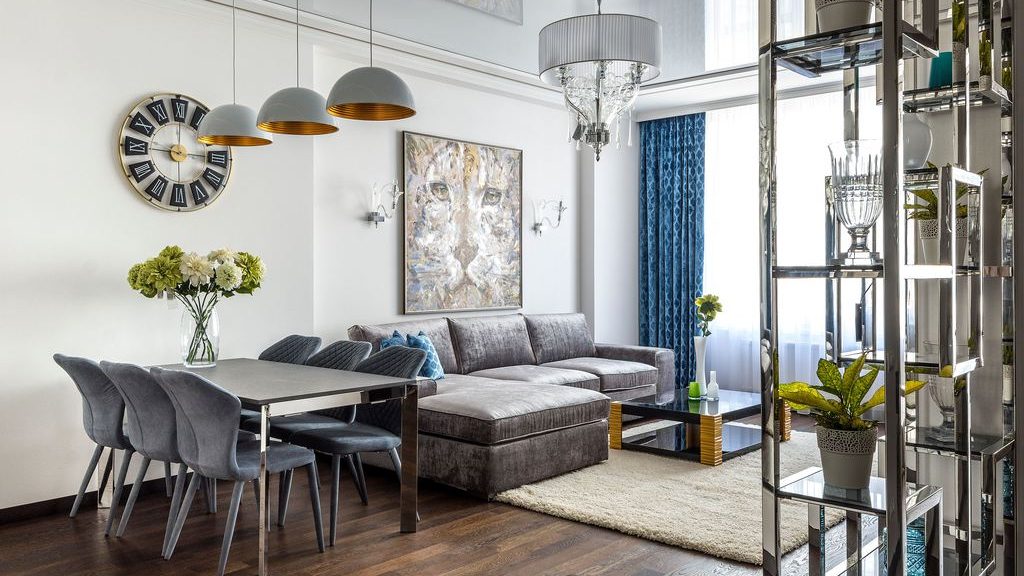 Open-concept studio living area with a gray sectional, dining table, large rug, blue curtains, and mirrored display shelving.