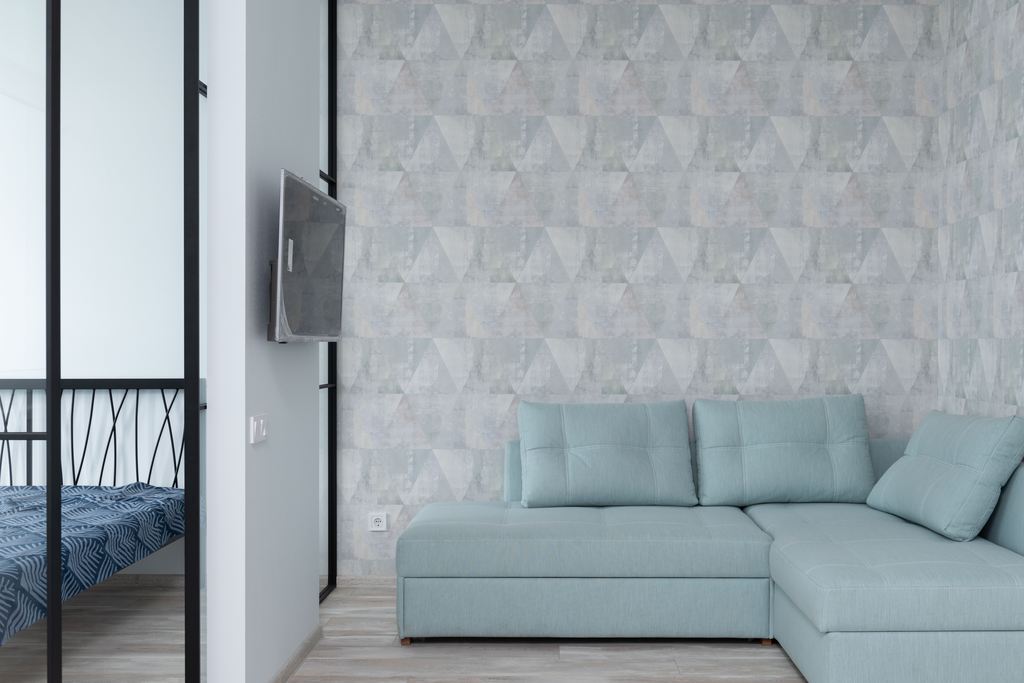 Wall-mounted TV beside a light blue sectional sofa in a compact modern apartment, with a geometric gray accent wall and an adjacent bedroom behind black-framed glass doors.