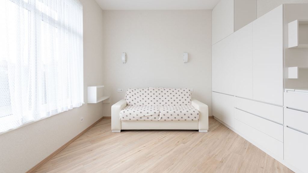Bright minimalist room with a small white sleeper sofa, sheer curtains, light wood flooring, and floor-to-ceiling white storage cabinets.