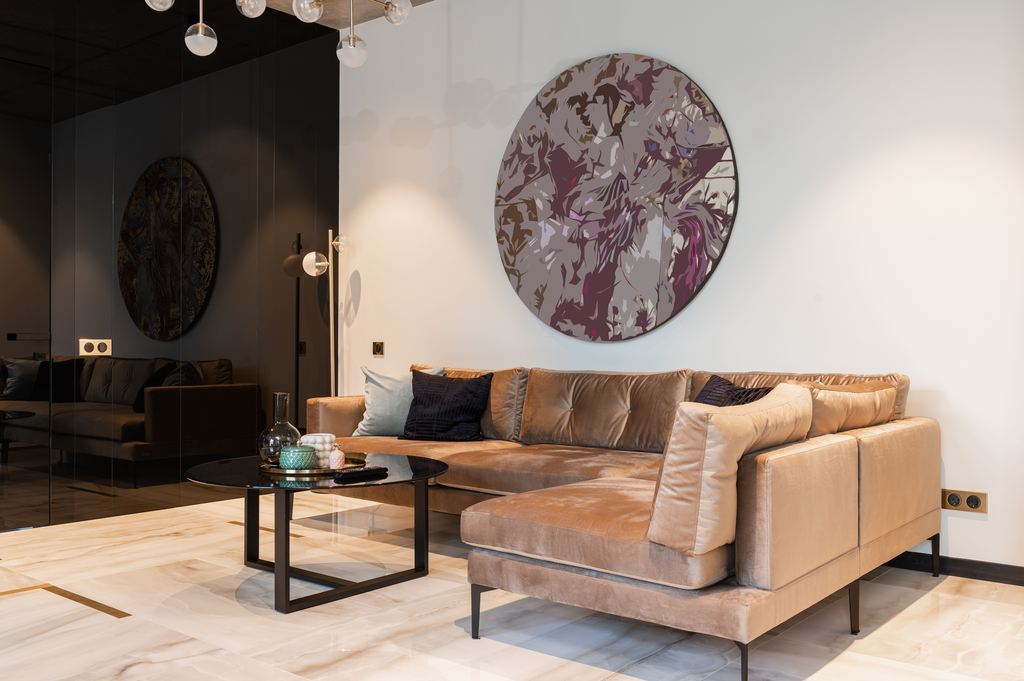Camel-brown sectional sofa with slim black legs in a modern living room with marble flooring, round wall art, and a black coffee table.