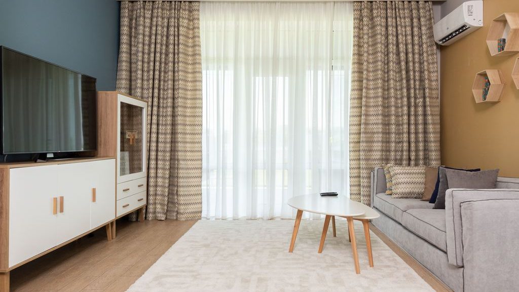 Modern living room with blue and mustard walls, a gray sofa, light wood furniture, patterned curtains, and a white area rug.