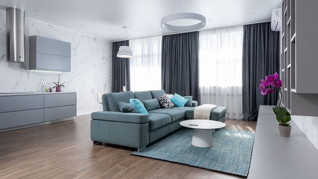Modern open-plan living room with a blue sectional sofa, layered teal pillows, a round white coffee table, gray cabinetry, and floor-to-ceiling curtains.