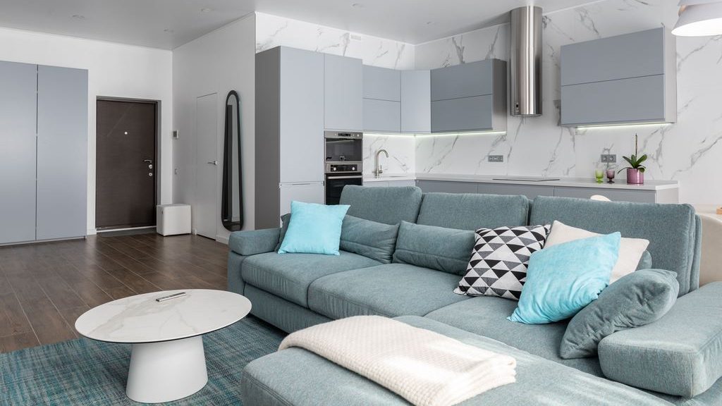 Open-plan studio apartment with a teal sectional sofa, round coffee table, and modern gray kitchen.